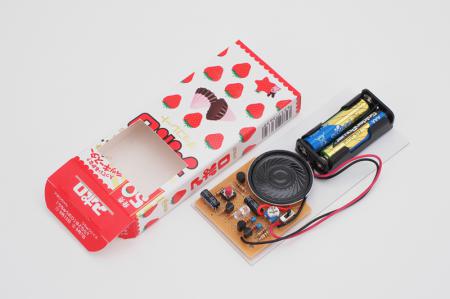 「アポロ」(明治)の箱に基板と電池ボックスを収めるよ。スピーカーやスイッチが出るように箱にはマドをあける。赤色LEDの光を細い線にするためには、箱のフタに細いスリットをあけている。