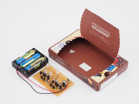 回路と電池は、箱(ここでは明治の「ALMOND　香ばしカカオニブ」の箱を使った)に両面テープを使って貼り付けている。箱は、光を当てるフィギュアなどが置けるスペースも考えて選んでね。